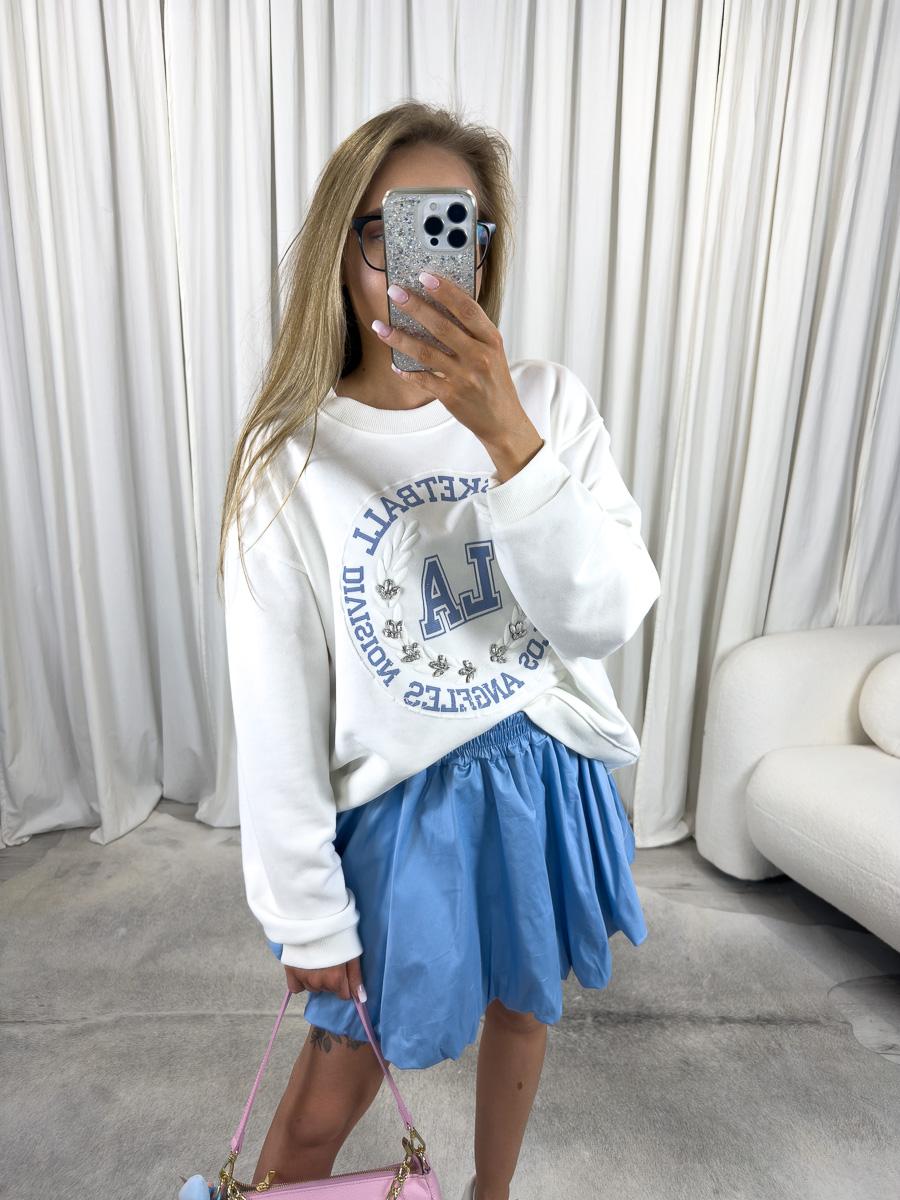 IMG_2717 Bluza oversize College biała z nadrukiem college, stylizacja z błękitną spódnicą, białymi sneakersami i różową torebką, zdjęcie produktowe na tle jasnych zasłon.