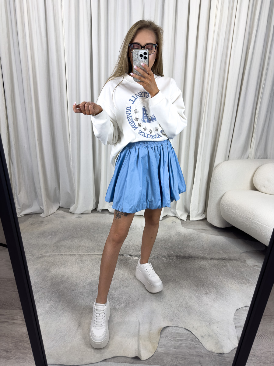 IMG_2724 Bluza oversize College biała z nadrukiem college, stylizacja z błękitną spódnicą, białymi sneakersami i różową torebką, zdjęcie produktowe na tle jasnych zasłon.