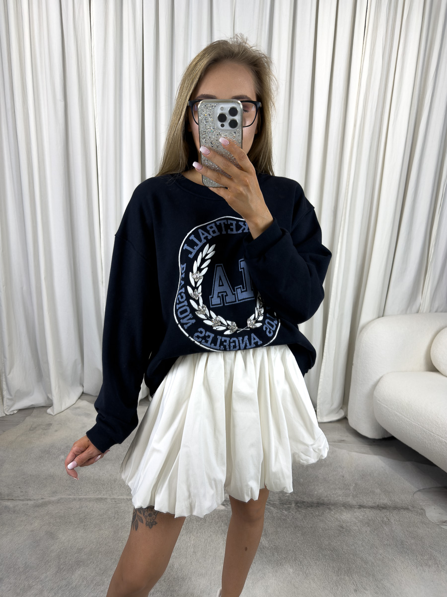 IMG_2730 Bluza oversize College granatowa z nadrukiem college, stylizacja z białą spódnicą i białymi sneakersami, zdjęcie produktowe na tle jasnych zasłon.