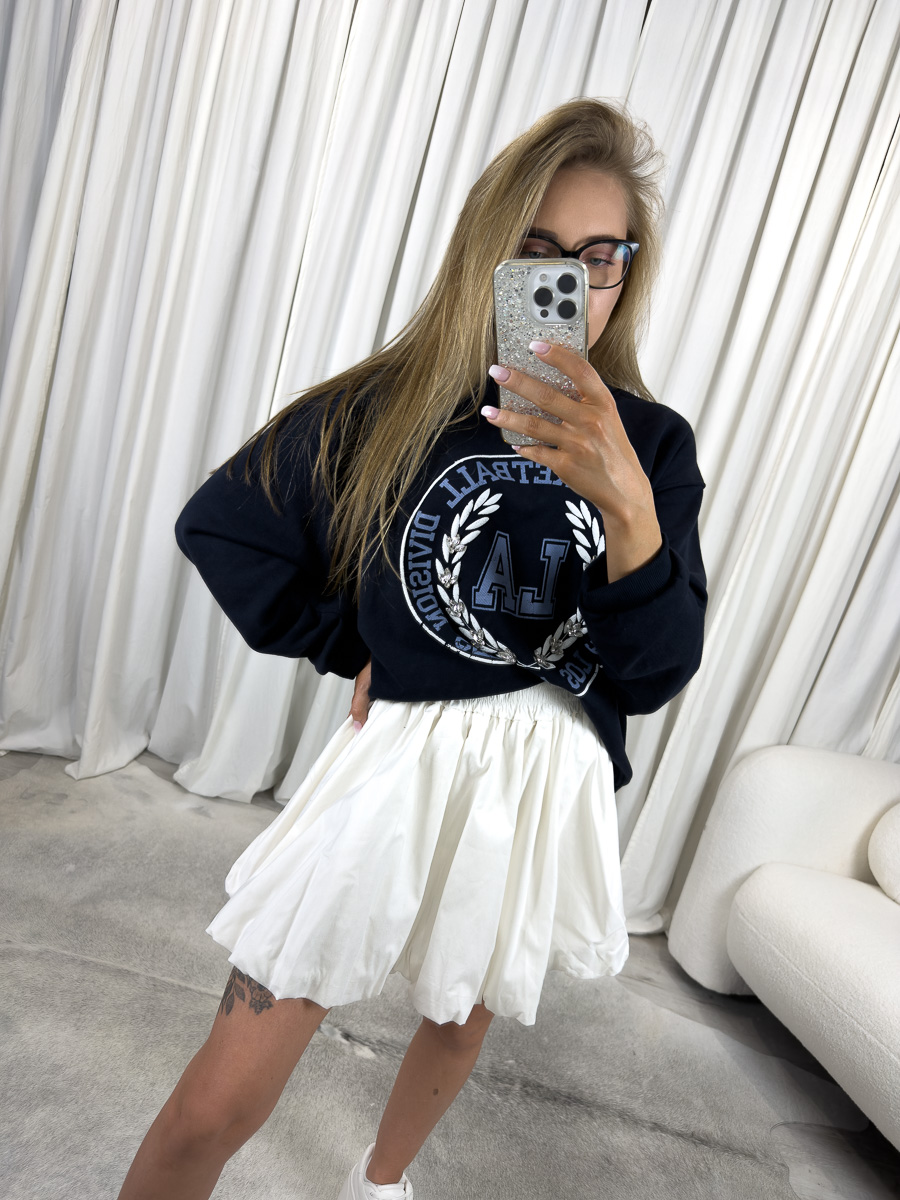 IMG_2739 Bluza oversize College granatowa z nadrukiem college, stylizacja z białą spódnicą i białymi sneakersami, zdjęcie produktowe na tle jasnych zasłon.