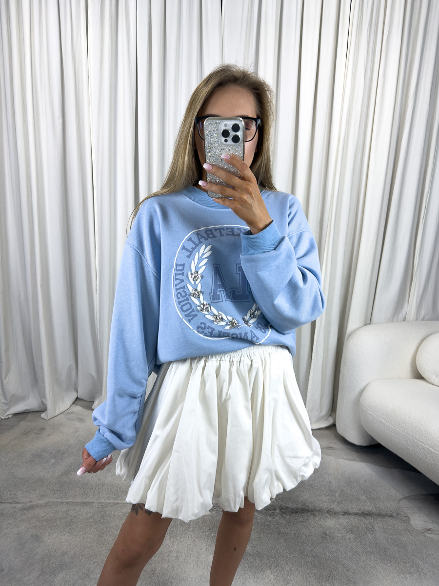 IMG_2757 Bluza oversize College błękitna z nadrukiem college, stylizacja z białą spódnicą i białymi sneakersami, zdjęcie produktowe na tle jasnych zasłon
