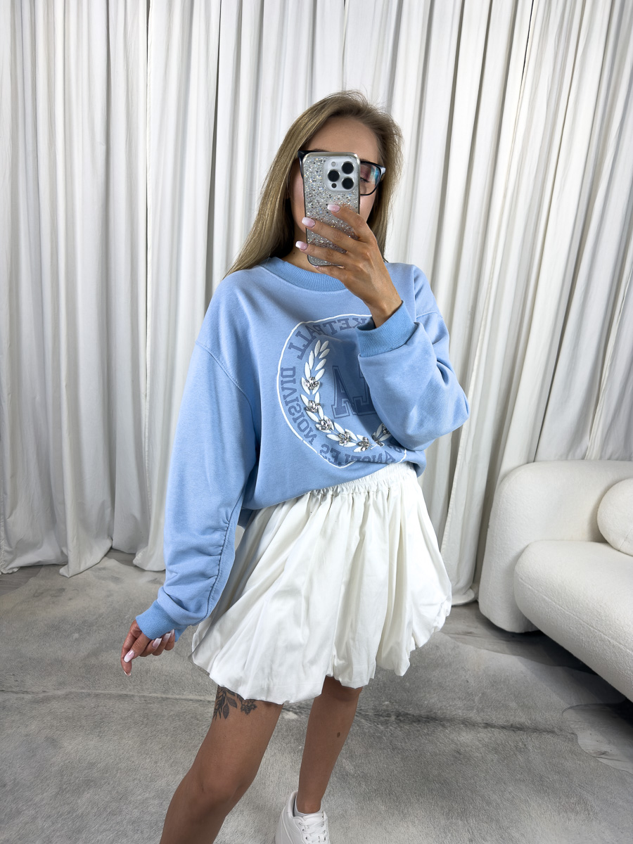 IMG_2760 Bluza oversize College błękitna z nadrukiem college, stylizacja z białą spódnicą i białymi sneakersami, zdjęcie produktowe na tle jasnych zasłon