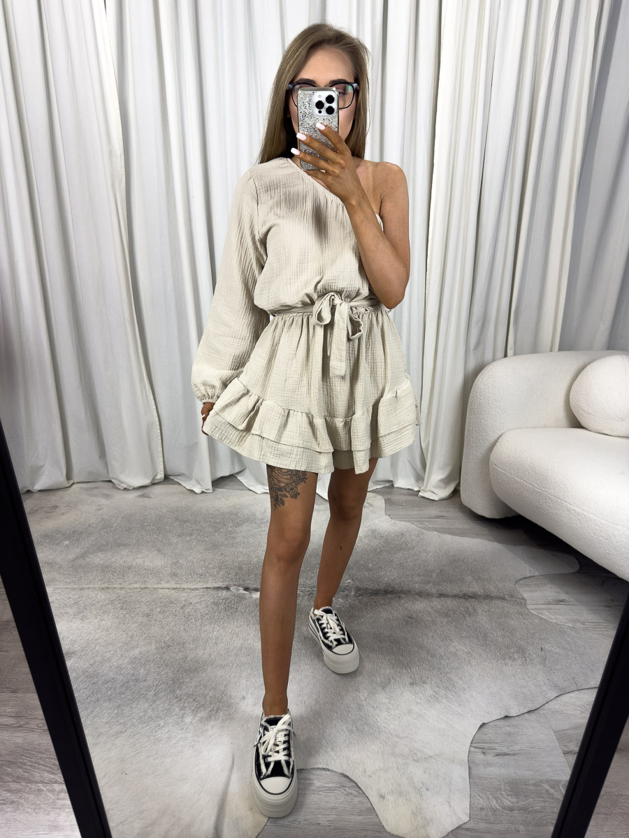muslinowy-kombinezon-girlish-bezowy Modelka w muślinowym kombinezonie Girlish z asymetrycznym ramieniem, falbankami i wiązaniem w talii. Zdjęcie produktowe sklepu internetowego z odzieżą damską.