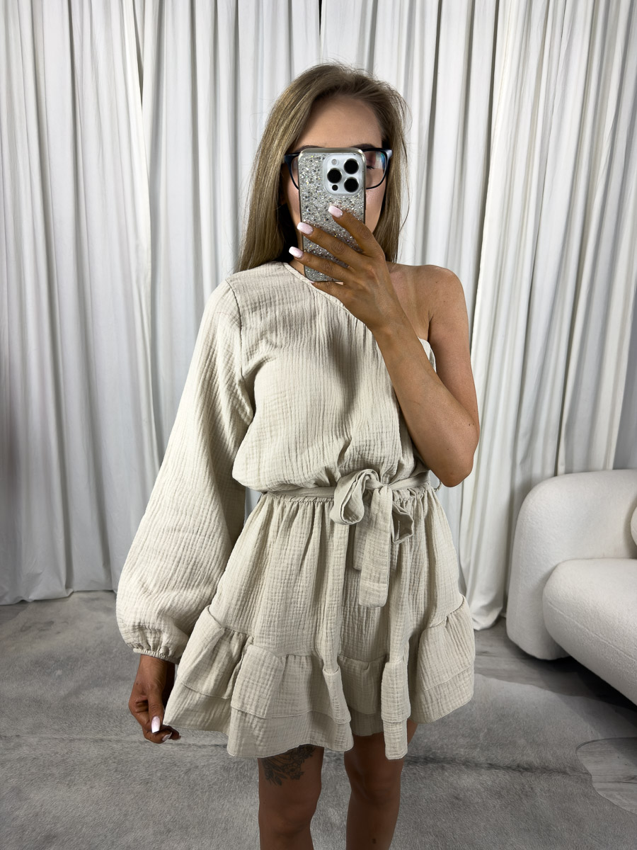muslinowy-kombinezon-girlish-bezowy Modelka w muślinowym kombinezonie Girlish z asymetrycznym ramieniem, falbankami i wiązaniem w talii. Zdjęcie produktowe sklepu internetowego z odzieżą damską.