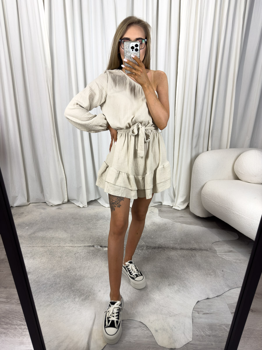 muslinowy-kombinezon-girlish-bezowy Modelka w muślinowym kombinezonie Girlish z asymetrycznym ramieniem, falbankami i wiązaniem w talii. Zdjęcie produktowe sklepu internetowego z odzieżą damską.