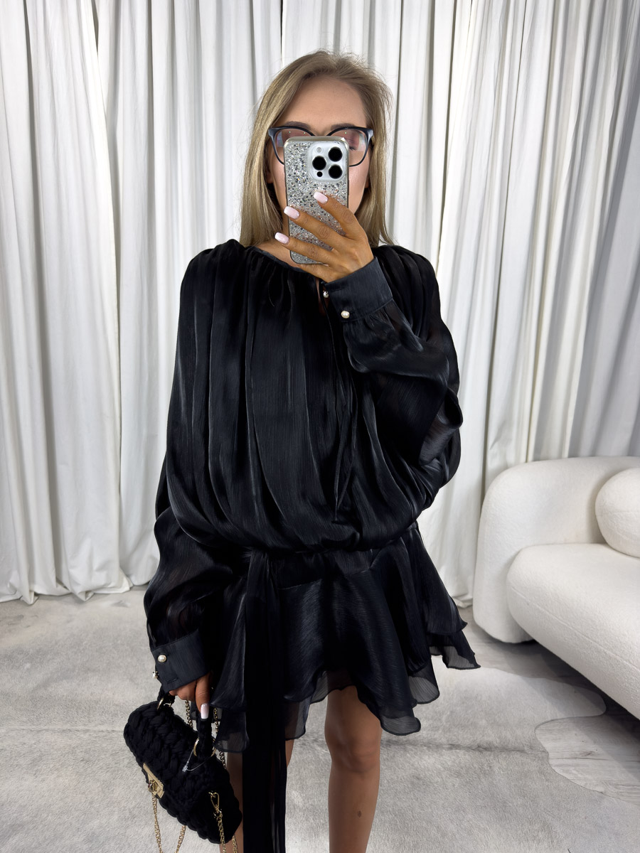 sukienka-mini-satin-czarna Modelka w czarnej sukience mini Satin z długimi, lejącymi rękawami, paskiem i falbaną na dole. Stylizacja z czarną torebką i szpilkami – zdjęcie produktowe do sklepu odzieżowego online.
