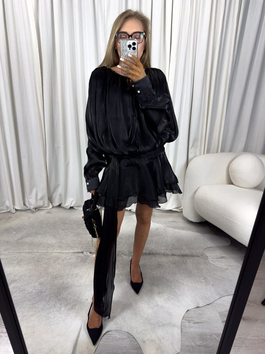sukienka-mini-satin-czarna Modelka w czarnej sukience mini Satin z długimi, lejącymi rękawami, paskiem i falbaną na dole. Stylizacja z czarną torebką i szpilkami – zdjęcie produktowe do sklepu odzieżowego online.
