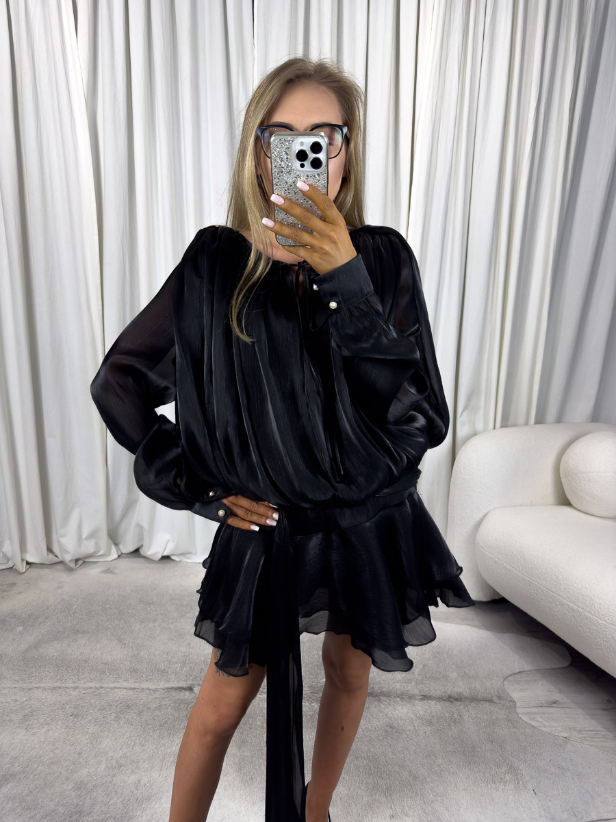 sukienka-mini-satin-czarna Modelka w czarnej sukience mini Satin z długimi, lejącymi rękawami, paskiem i falbaną na dole. Stylizacja z czarną torebką i szpilkami – zdjęcie produktowe do sklepu odzieżowego online.